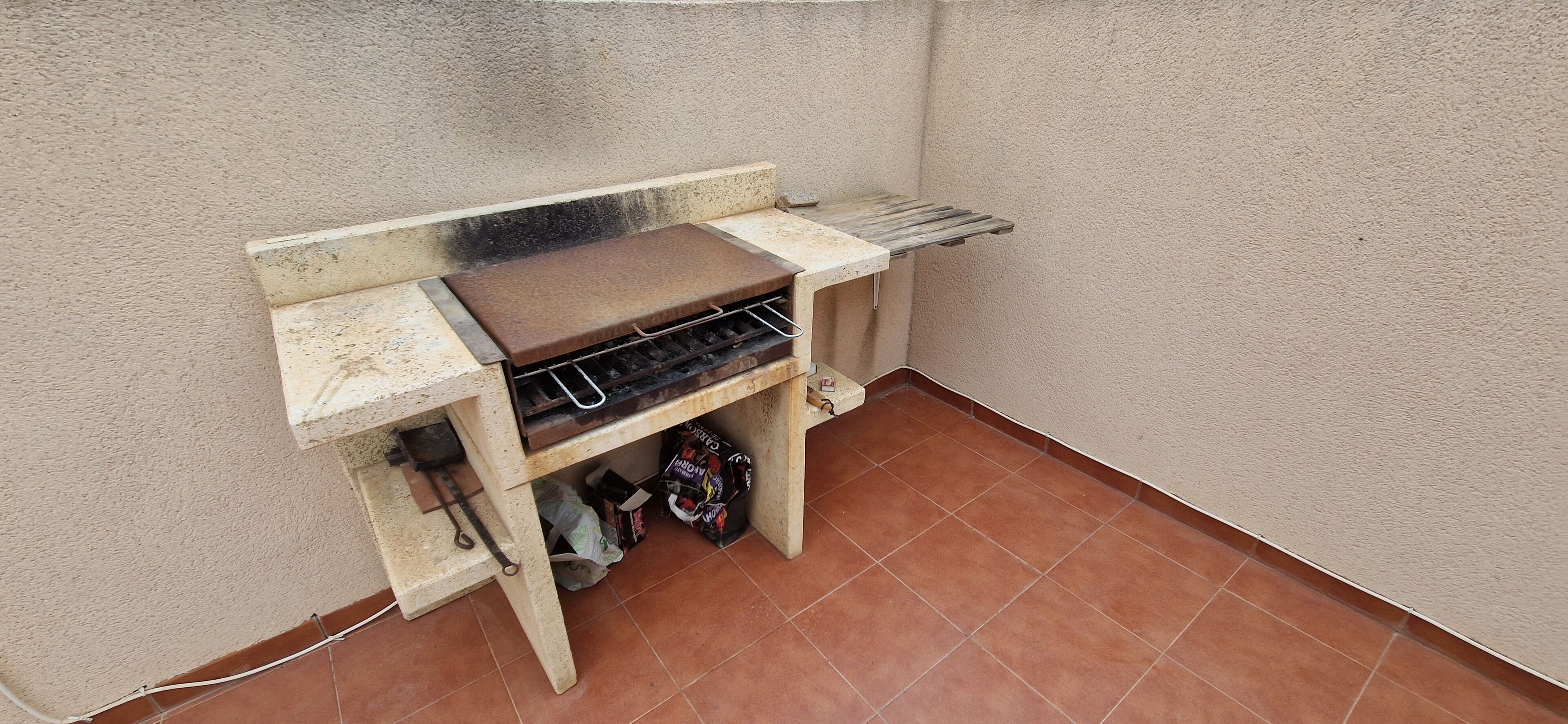 Un petit espace extérieur aménagé en coin barbecue, avec un gril en métal, un banc en bois et un sol carrelé.