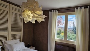 A bedroom with a bed, a nightstand, a window with curtains, and a view of trees and hills. - Daniela's House in Peace and Comfort (Sarlat-La-Canéda)
