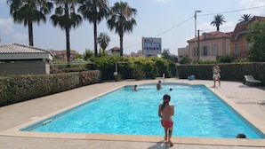 A swimming pool with clear blue water, surrounded by palm trees and a hedge.