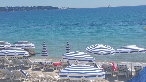 A beach with rows of sun loungers and striped umbrellas, a clear view of the sea, and a distant ship.