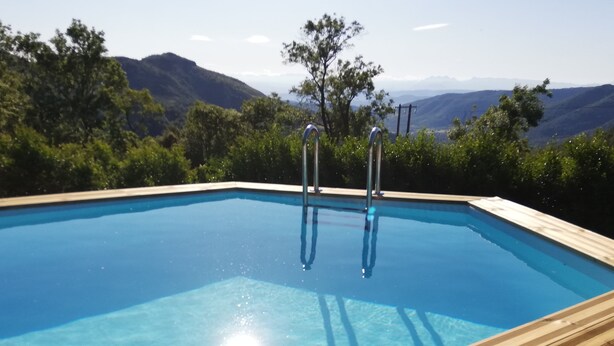 Una piscina rectangular de madera con escaleras metálicas, rodeada de vegetación exuberante y montañas al fondo.