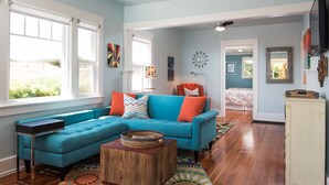 A spacious living room with a blue sofa, wooden coffee table, and a view through large windows. - Ocean Beach Sunset Cottage - Steps to Shoreline (San Diego)