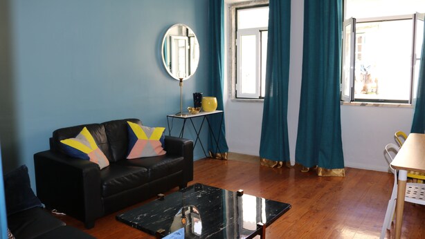 Una sala de estar con un sofá de cuero negro, un espejo redondo, una mesita y cortinas color turquesa.