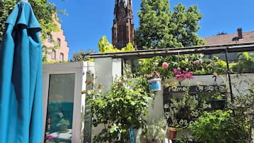 Un jardin sur le toit, agrémenté de plantes et de fleurs en pot, avec en arrière-plan le clocher d’une église et un ciel bleu dégagé.