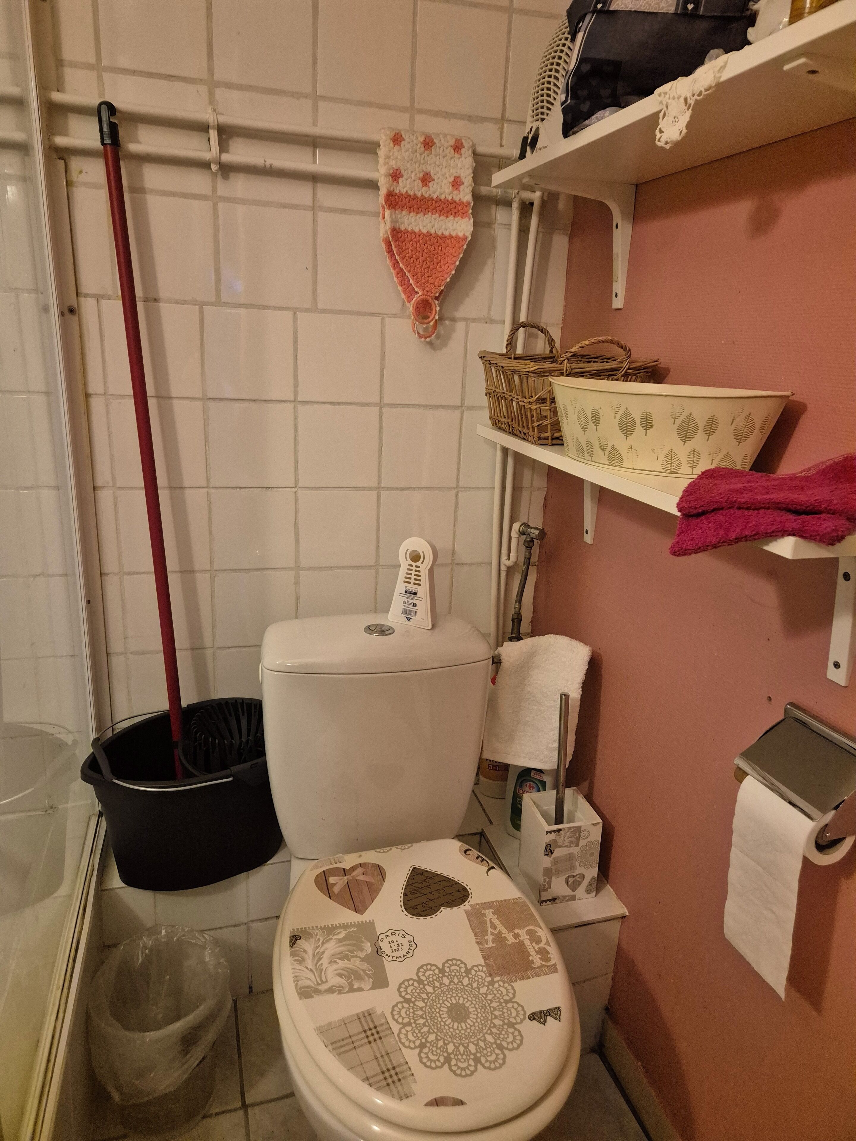 A bathroom with a toilet, a bidet, a mop, a trash bin, and a shelf with a basket and a pink towel.