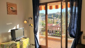 A room with a television on a table, a window with curtains, and a view of a residential area. - Residence Provençale Pool Parking WIFI 2 kms Sea Villeneuve Loubet 2 Rooms (Villeneuve-Loubet)
