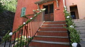 Un immeuble de deux étages avec une façade rouge, des volets verts et un escalier menant à l’entrée.
