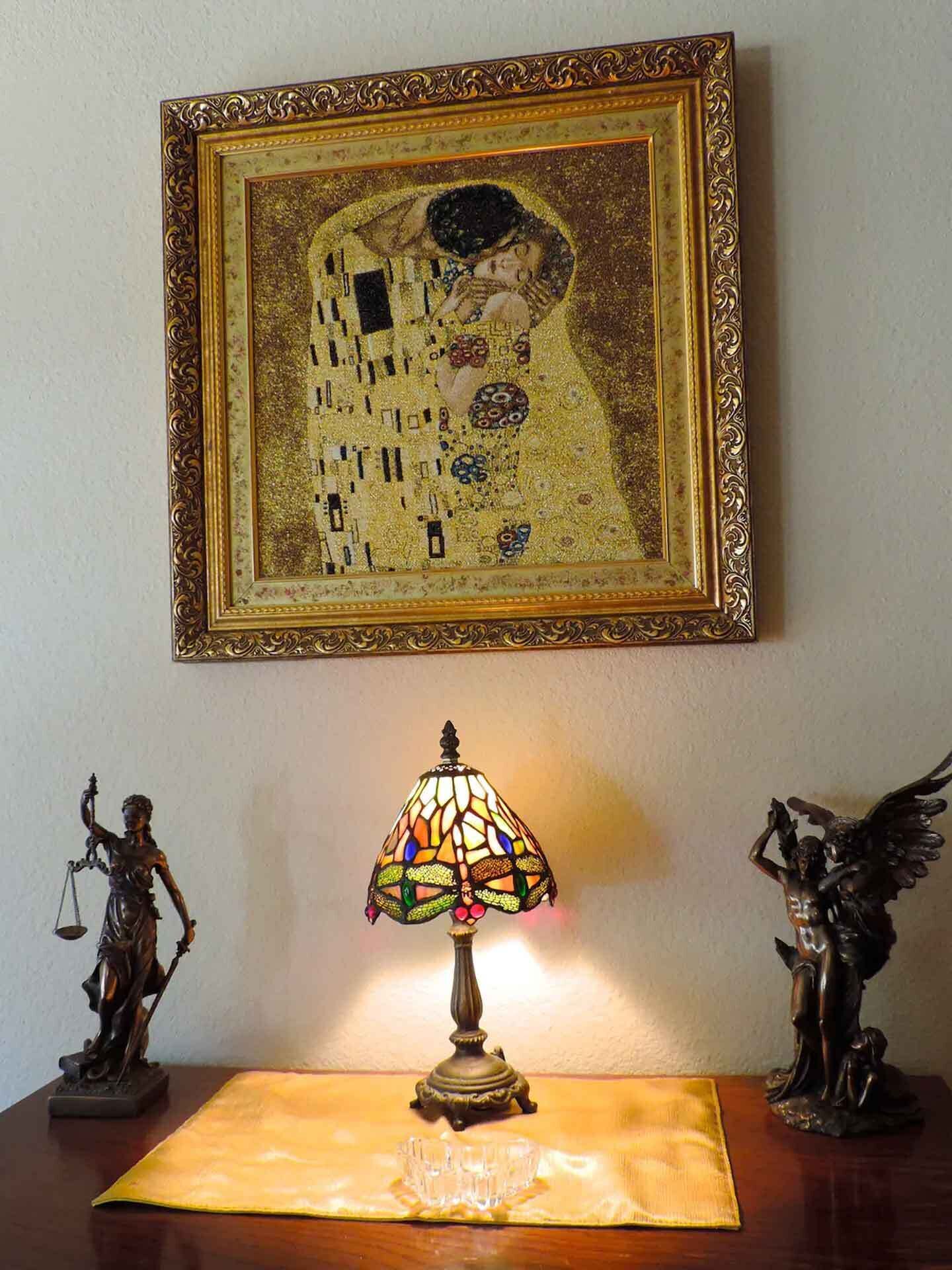 A framed Gustav Klimt painting, a stained glass lamp, and two bronze statues on a wooden table.