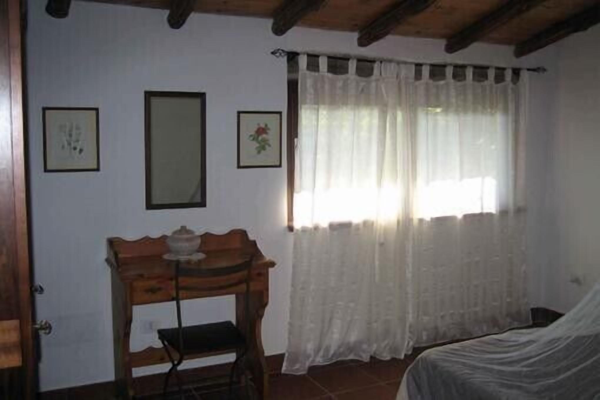 A bedroom with a bed, a wooden nightstand, a chair, and a window with sheer curtains.