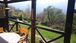 Terraza de madera con mesa y sillas, con vista a un jardín y montañas lejanas.