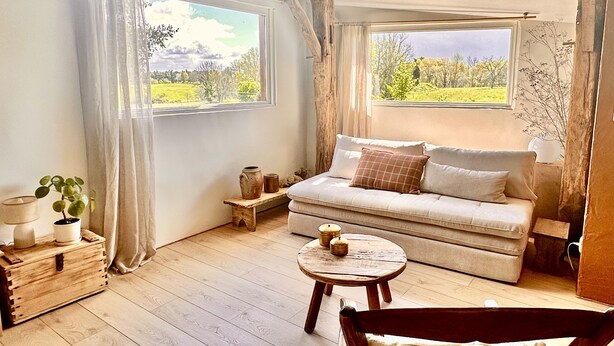 A cozy room with a wooden floor, a sofa, a small round table, and a view of a green landscape through large windows. - The small sheepfold in the Perigord (Villefranche-Du-Périgord)