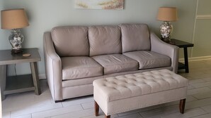 A living room with a beige sofa, a matching ottoman, two side tables with lamps, and a beach-themed painting on the wall. - Beautiful White Sand Gulf Beaches-Orange Beach, Alabama (Orange Beach)