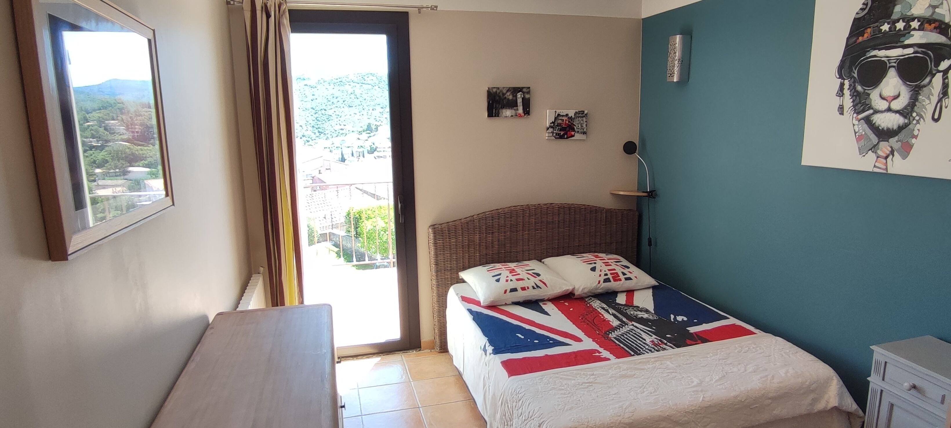 Un dormitorio con una cama que presenta un diseño con la bandera Union Jack, un cabecero de mimbre y vistas a un paisaje montañoso a través de una puerta corrediza.
