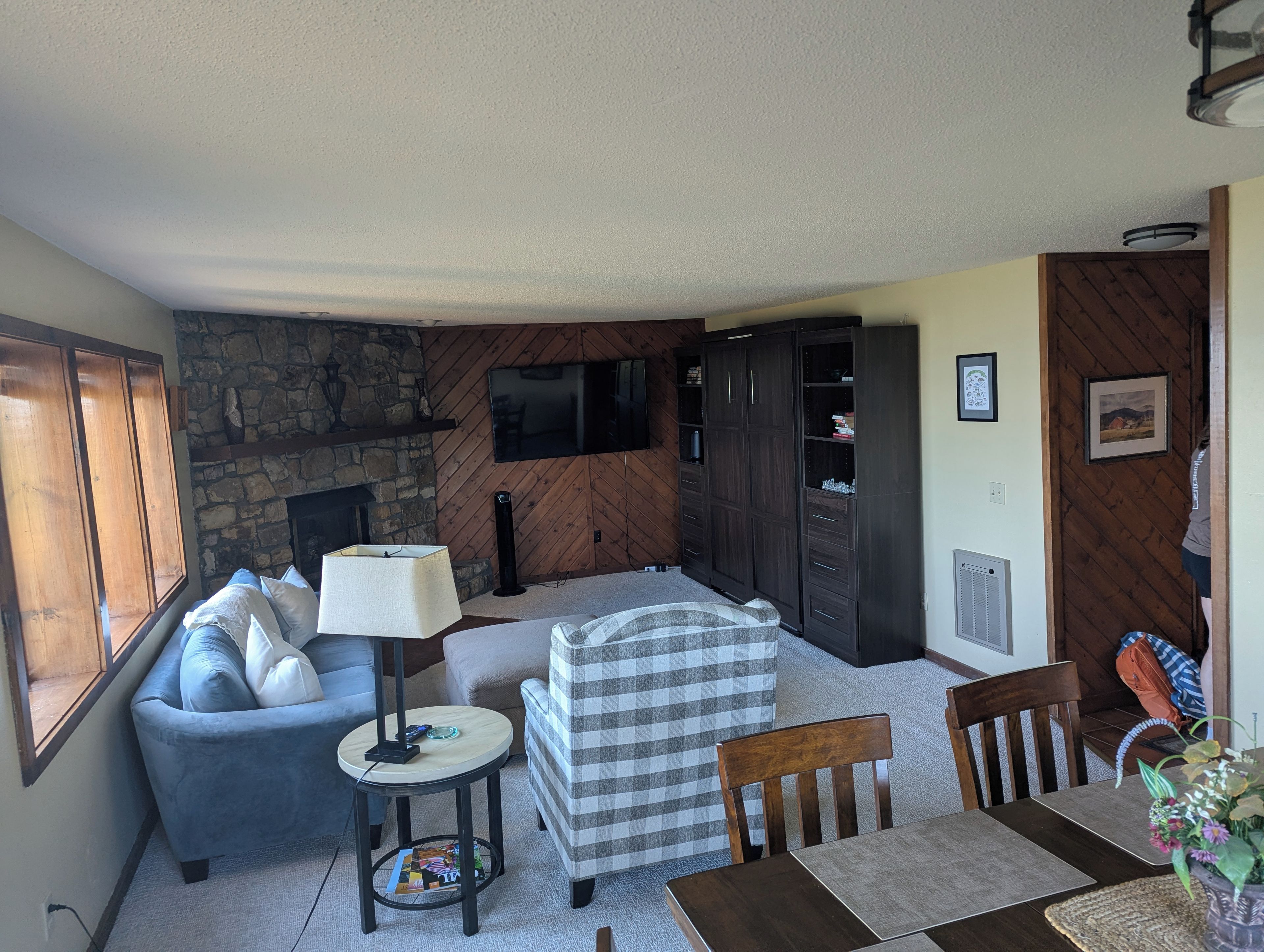 Ein Wohnzimmer mit Kamin aus Stein, einem an der Wand montierten Fernseher, einer Couch, einem gestreiften Ohrensessel, einem Esstisch mit Stühlen und einer Lampe.