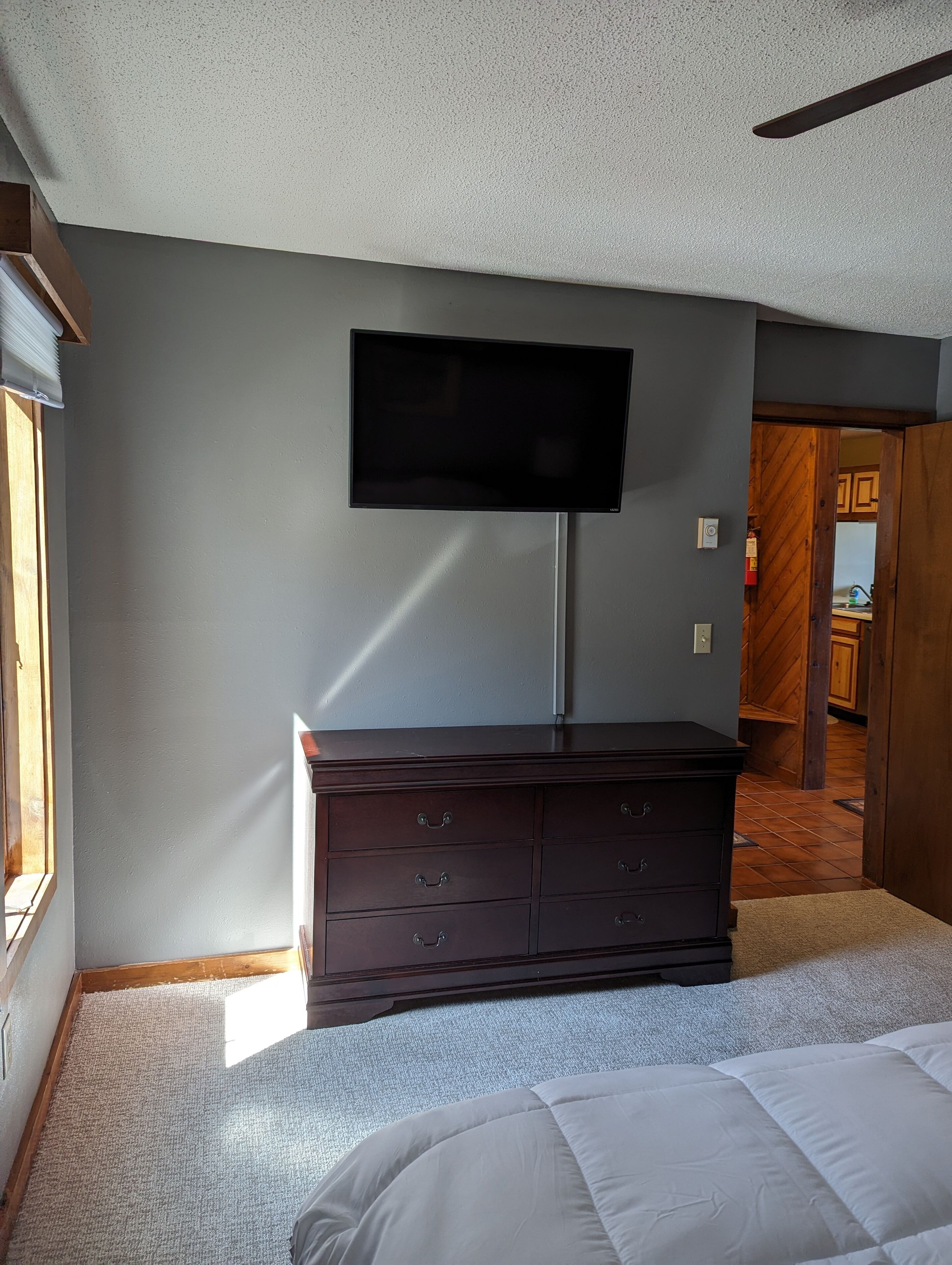 A bedroom with a large bed, a dresser, a flat-screen TV mounted on the wall, and a window providing natural light.