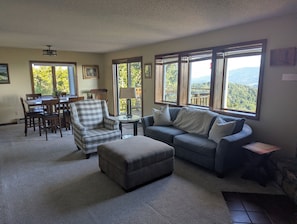 A living room with a sofa, ottoman, and armchair, a dining table with chairs, and large windows with a view of trees and hills. - Fantastic view! Condo located on the Crest of Sugar Mountain (Banner Elk)