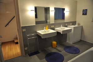 Une salle de bain moderne avec deux lavabos, un miroir et des toilettes. Il y a deux tapis bleus sur le sol.