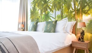 Ein Hotelzimmer mit Bett, Kissen, Nachttisch, Lampe und einer Wandmalerei von Palmen.