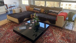 A modern living room with a sectional sofa, a glass coffee table with decorative items, a patterned rug, and a view of the outdoors through large windows.