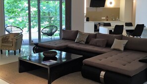 Un salon moderne avec un canapé d’angle, une table basse en verre et une vue sur l’extérieur grâce à de grandes fenêtres.