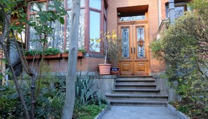 A two-story building with a wooden front door, a brick facade, and a small front garden with steps leading to the entrance.