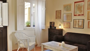 A living room with a white wicker chair, a dark brown sofa, a wooden coffee table, and a window with white curtains.