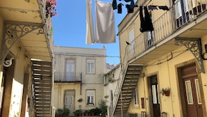 A narrow street with buildings on either side, balconies with clothes hanging, and potted plants.