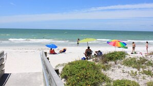 Una playa con gente relajándose bajo sombrillas, un paseo marítimo de madera y vistas al océano.