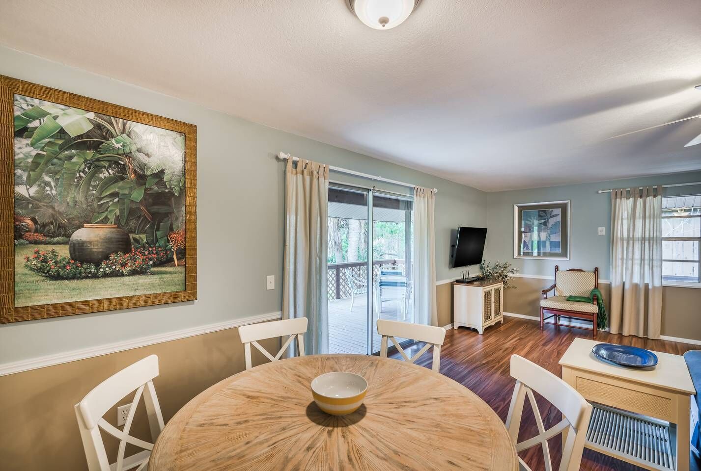 Un coin repas comprenant une table ronde, des chaises, un téléviseur et un tableau représentant un paysage tropical.