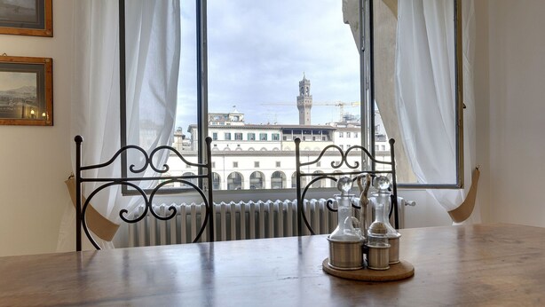 A room with a table, two chairs, a window with curtains, a radiator, and a view of buildings and a tower. - Bardi Apartment, Stunning View Of Ponte Vecchio! (Florence)