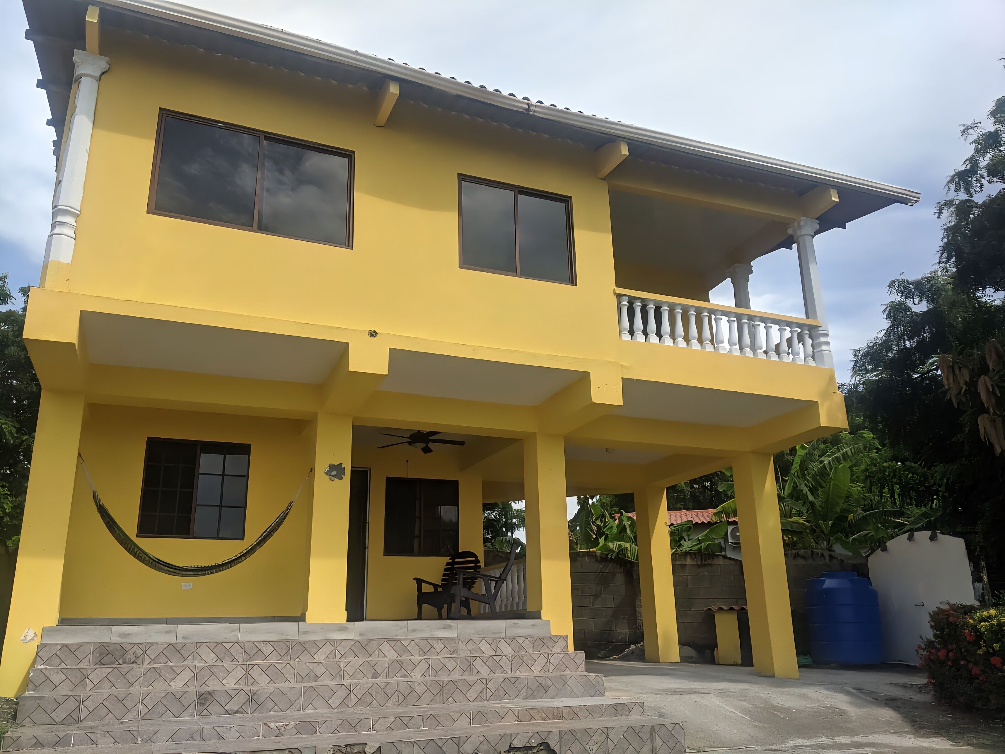 Un edificio giallo di due piani con un balcone, una scala che conduce all'ingresso e una piccola zona coperta con una ventola a soffitto.