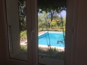 Une vue à travers une fenêtre sur une piscine, entourée de verdure et agrémentée d’un banc en bois.