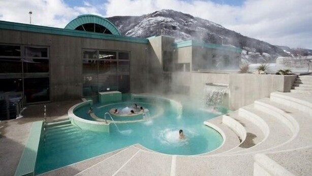 Un área de spa con hidromasaje y piscina, rodeado por un edificio con amplios ventanales y una montaña al fondo.