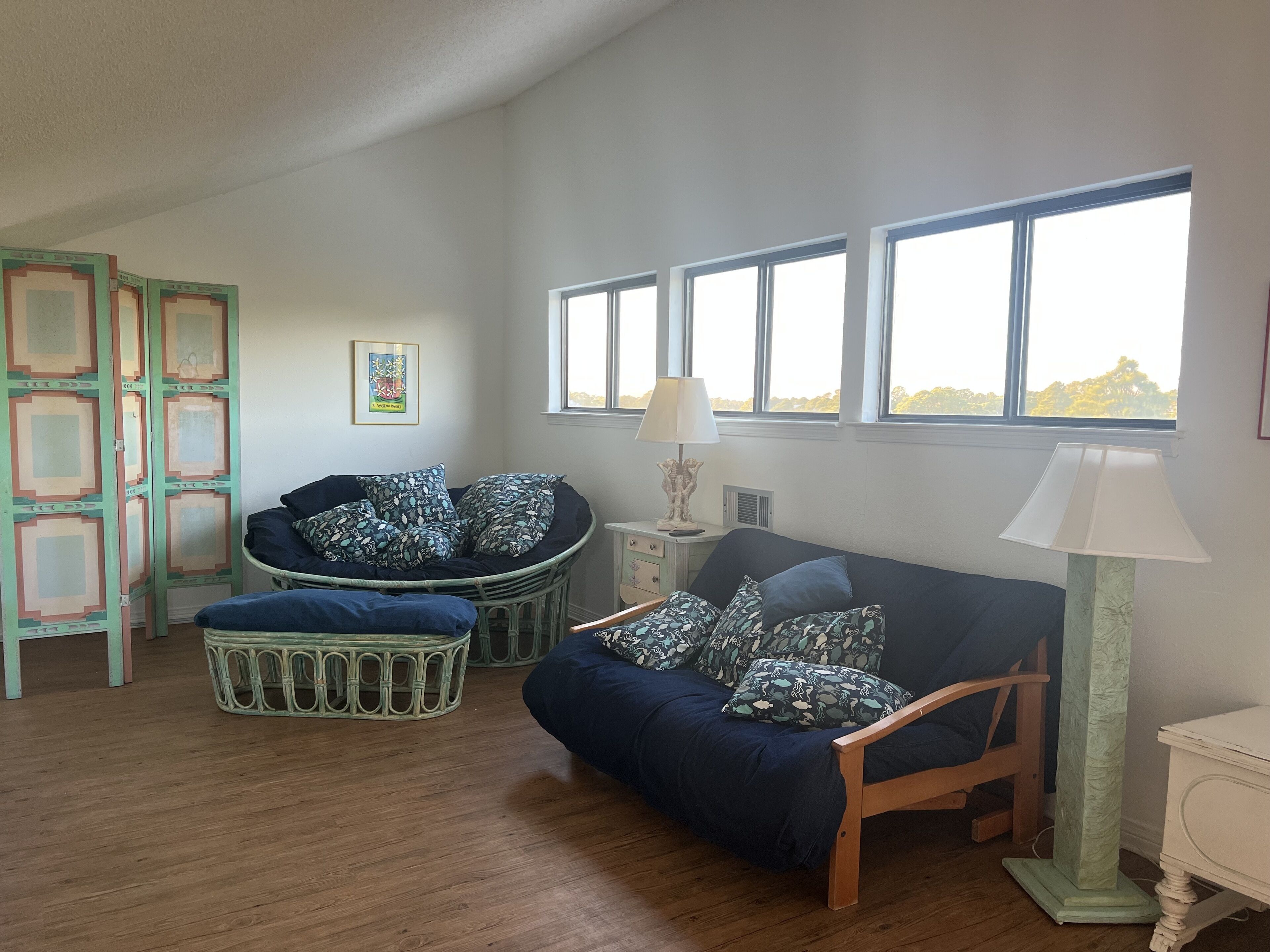 Een gezellige kamer met een donkerblauwe bank, een bijpassend voetenbankje, een houten stoel, een klein bijzettafeltje met een lamp, en een raam met uitzicht op bomen.