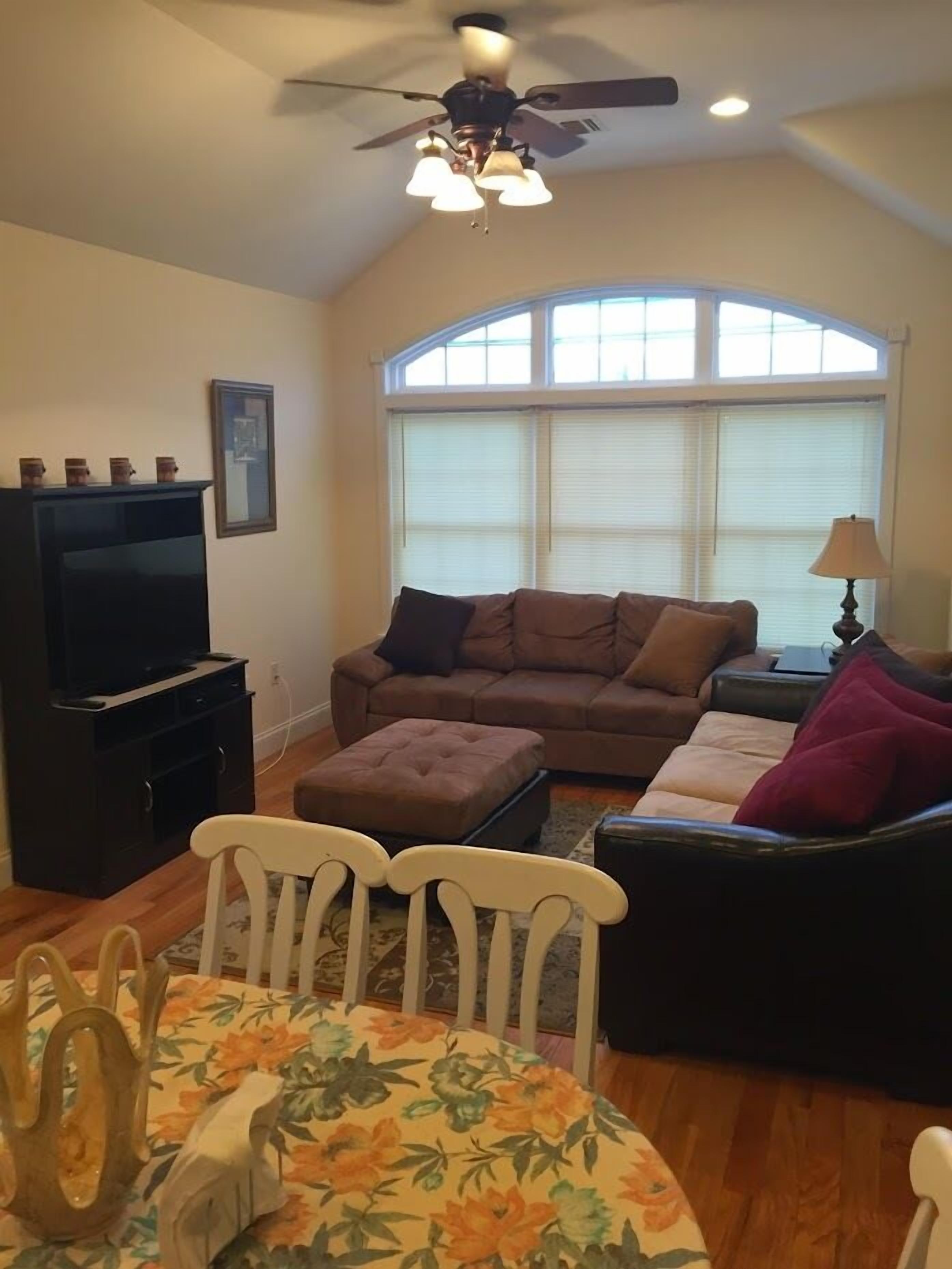 A living room with a brown sofa, a television, a ceiling fan, and a dining table with chairs.