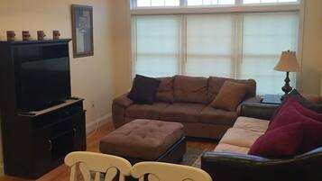 A living room with a brown sofa, a television, a ceiling fan, and a dining table with chairs.