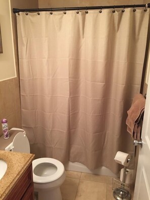 Un baño con cortina de ducha color beige, inodoro blanco, encimera color beige y un perchero.