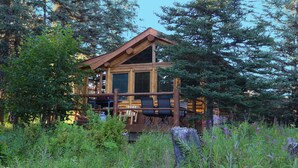 A wooden cabin with a deck, surrounded by dense forest. - Moose - Unique Alaskan log home in trees & wildflowers on 14-acre homestead. (Anchor Point)
