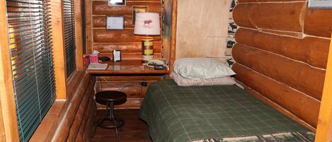 A small cabin room with a bed, a desk with a lamp, and a chair.
