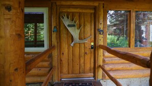 A wooden cabin door with a moose antler decoration, a window with a view of trees, and a wooden railing. - Moose - Unique Alaskan log home in trees & wildflowers on 14-acre homestead. (Anchor Point)