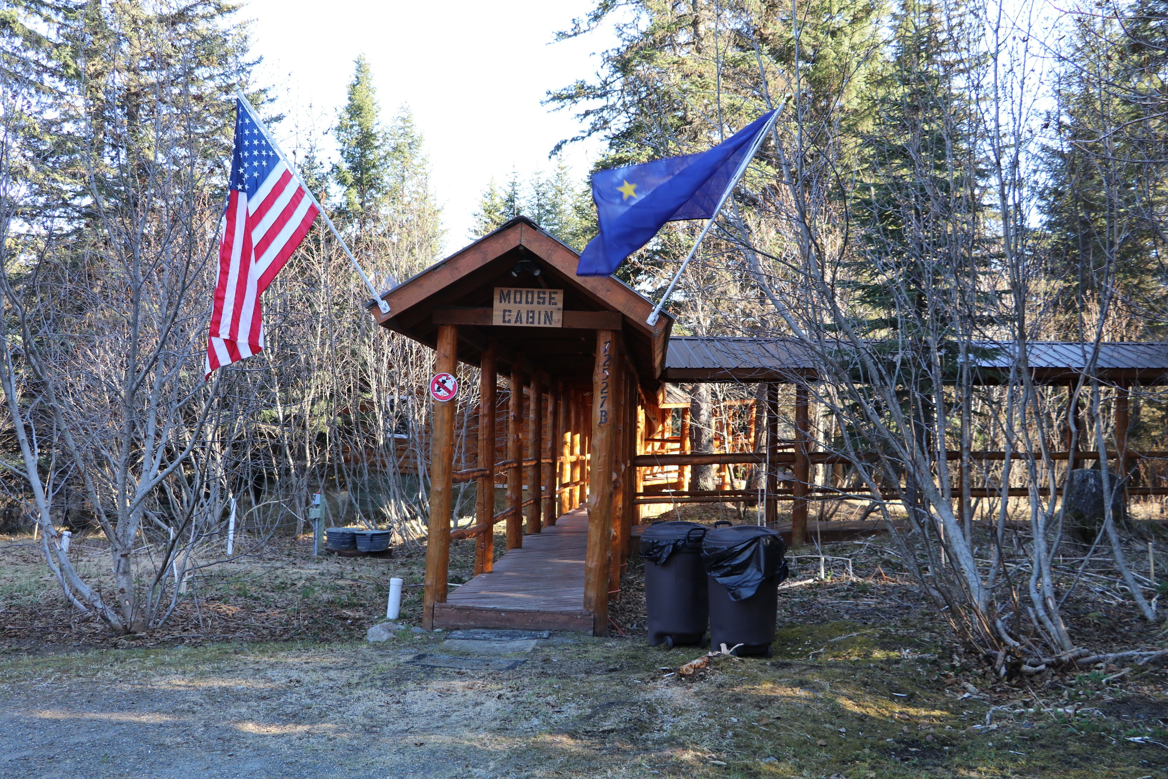 Une cabane en bois avec une pancarte indiquant « CABANE À ORIGNETS » et deux drapeaux, un américain et un européen, située dans une zone boisée.