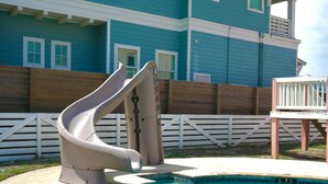 Una piscina con tobogán, una terraza de madera y una casa con revestimiento azul y detalles blancos.