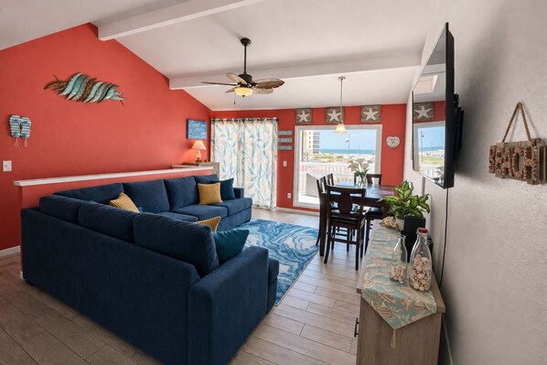 A living room with a blue sofa, a dining table with chairs, a ceiling fan, and a view of the ocean through a large window.