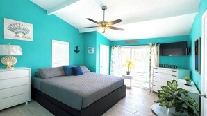 Un dormitorio con cama, ventilador de techo, televisor, planta y ventana con cortinas.