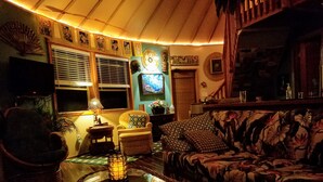 A cozy living room with a sofa, armchair, and a glass coffee table. There is a lamp on a side table, a wall-mounted shelf with decorative items, and a window with blinds. -  New Round House With Pool 180 Degree Ocean View 13% discount for 7 nights  (Captain Cook)