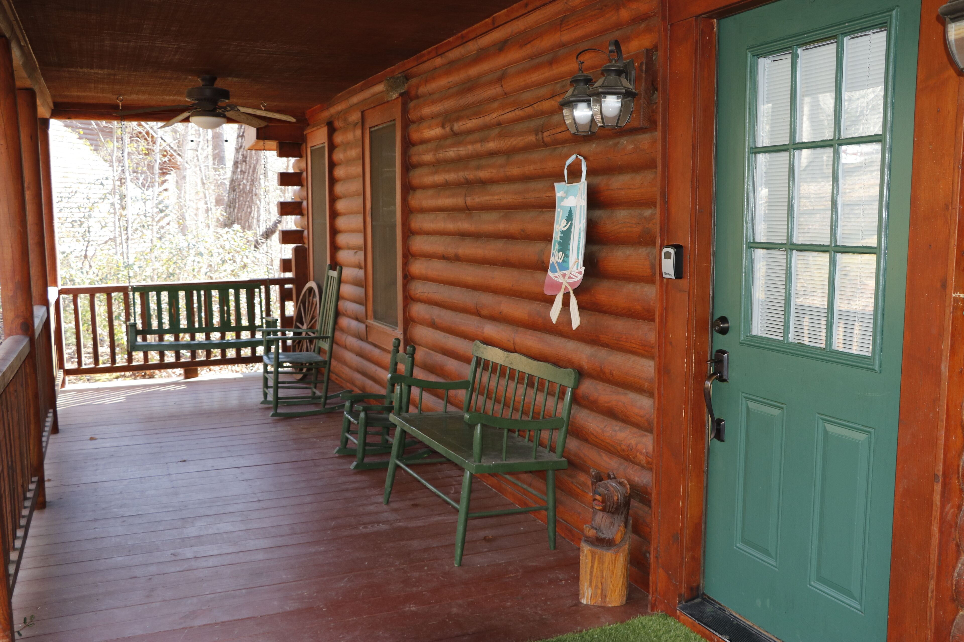 A wooden cabin with a porch, green benches, a door with a window, and a lantern.
