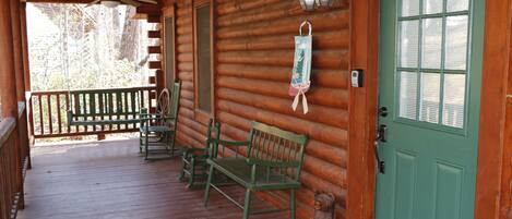 A wooden cabin with a porch, green benches, a door with a window, and a lantern.