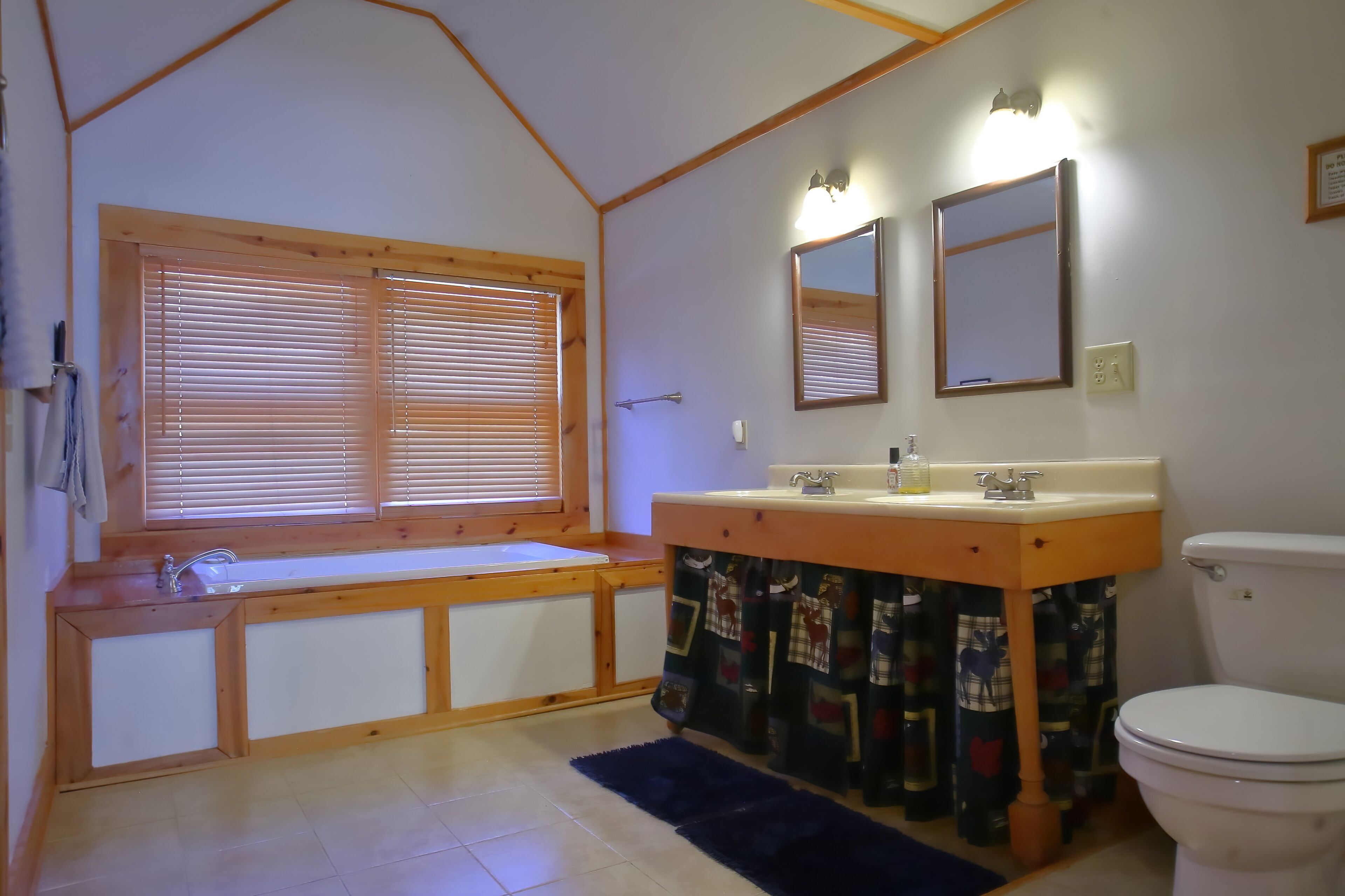 Un baño con tocador de madera, dos espejos, inodoro y ventana con persianas.