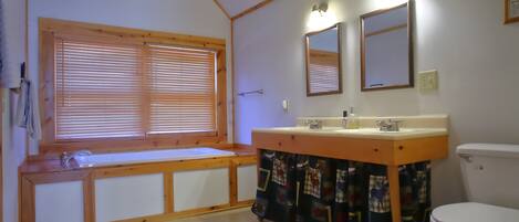 Un baño con tocador de madera, dos espejos, inodoro y ventana con persianas.