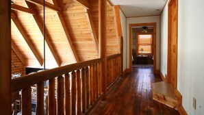 Interior em madeira com teto inclinado, escada de madeira e corredor que dá para outro quarto.